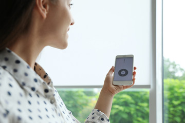 Woman using smart home application on phone to control window blinds indoors