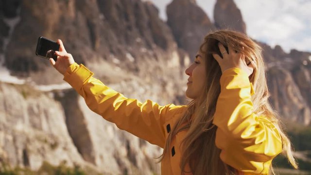 Young cheerful woman in a yellow raincoat taking selfie on beautiful mountains background during sunny day, slow motion