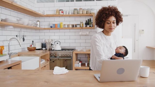 Multi-Tasking Mother Holds Sleeping Baby Son Whilst Working On Laptop In Kitchen