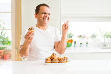 Middle age man eating chocolate chip muffins at home very happy pointing with hand and finger to the side