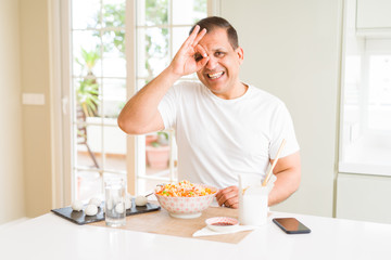 Middle age man eating asian food with chopsticks at home doing ok gesture with hand smiling, eye looking through fingers with happy face.