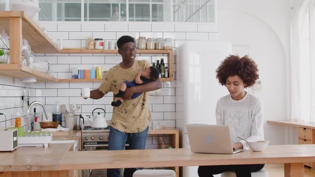 Busy Family In Kitchen At Breakfast With Mother Working On Laptop And Father Caring For Baby Son