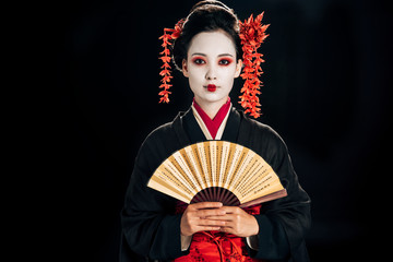 Obraz premium geisha in black kimono with red flowers in hair holding traditional asian hand fan isolated on black