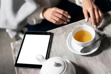 mock up tablet lies on table in cafe next to cup of tea and teapot. At the table sits girl and reads from screen. Copy space.