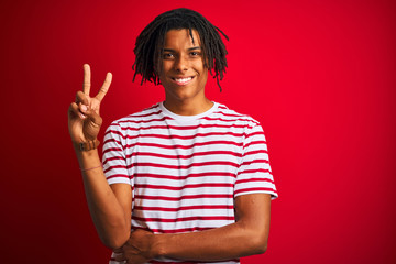 Young afro man with dreadlocks wearing striped t-shirt standing over isolated red background smiling looking to the camera showing fingers doing victory sign. Number two.