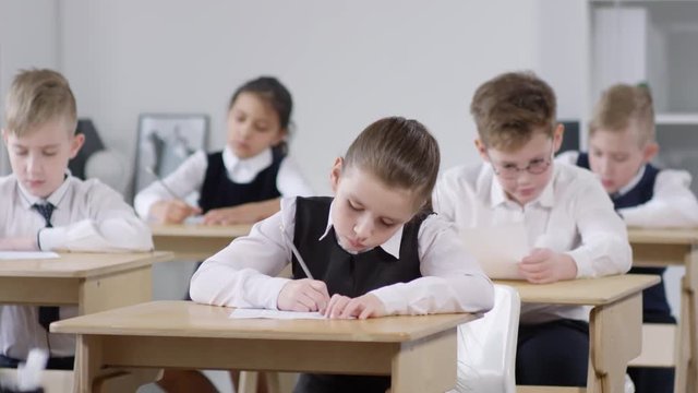 Medium tracking shot of group of six preteen Caucasian schoolchildren, wearing uniform white shirts and pinafores, sitting at their desks and writing in test answer sheets