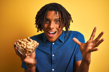 Afro american man with dreadlocks eating peanuts over isolated yellow background very happy and excited, winner expression celebrating victory screaming with big smile and raised hands