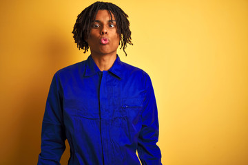 Obraz premium Afro worker man with dreadlocks wearing mechanic uniform over isolated yellow background making fish face with lips, crazy and comical gesture. Funny expression.