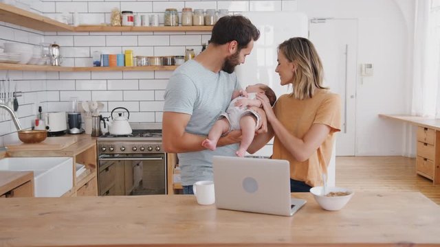 Busy Family In Kitchen At Breakfast With Mother Working On Laptop And Father Caring For Baby Son