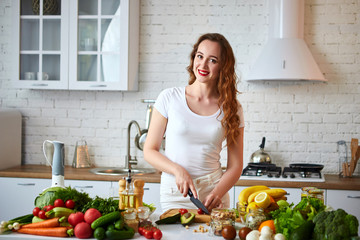 Young happy woman cutting tomatoes for making salad in the beautiful kitchen with green fresh ingredients indoors. Healthy food and Dieting concept. Loosing Weight