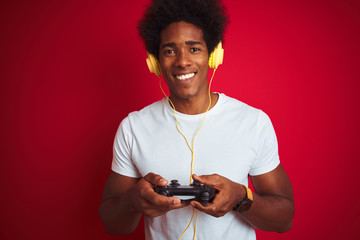Afro gamer man playing video game using joystick headphones over isolated red background with a happy face standing and smiling with a confident smile showing teeth © Krakenimages.com