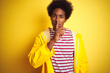 African american man with afro hair wearing rain coat standing over isolated yellow background asking to be quiet with finger on lips. Silence and secret concept.