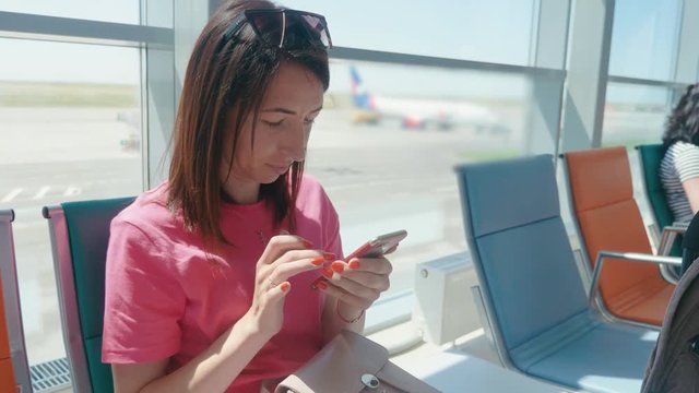 A young beautiful girl sits in the airport waiting room and uses a mobile phone. Woman buying e-ticket, making hotel reservations and checking in online.