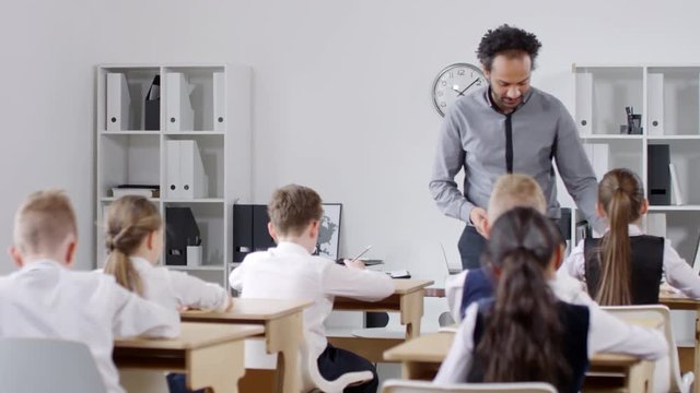 Full shot of cheerful Afro-American male teacher standing up from his desk, walking between rows of desks and collecting test answer sheets from elementary school pupils, while talking and joking