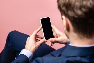 back view of businessman sitting and holding smartphone with blank screen isolated on pink