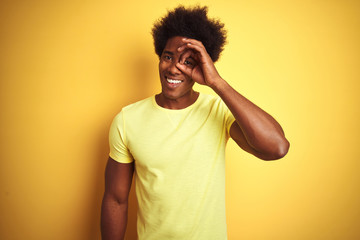 African american man with afro hair wearing t-shirt standing over isolated yellow background doing ok gesture with hand smiling, eye looking through fingers with happy face.