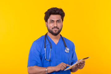 Cute Indian doctor in a blue robe with a stethoscope looking at a tablet based on a yellow background. Concept online consultation