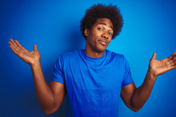 Fototapeta na wymiar African american man with afro hair wearing t-shirt standing over isolated blue background clueless and confused expression with arms and hands raised. Doubt concept.
