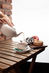 Woman is pouring tea for a tea party with doughnuts: strawberry, oreo and chocolate glazed, served on a brick wall background