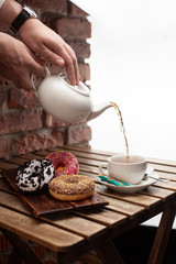 Woman is pouring tea for a tea party with doughnuts: strawberry, oreo and chocolate glazed, served on a brick wall background