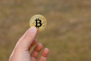 Man fingers are holding a big round golden bitcoin coin in front of yellow green pattern.