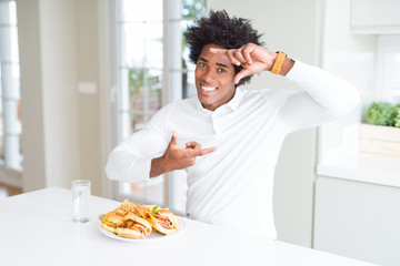 Fototapeta na wymiar African American hungry man eating hamburger for lunch smiling making frame with hands and fingers with happy face. Creativity and photography concept.