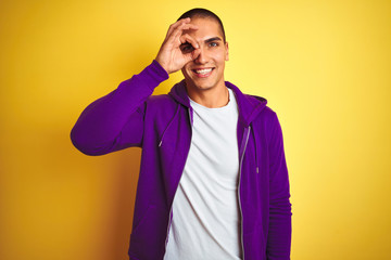 Young handsome man wearing purple sweatshirt over yellow isolated background doing ok gesture with hand smiling, eye looking through fingers with happy face.