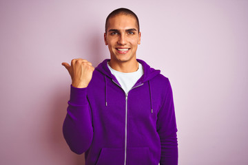 Young handsome man wearing purple sweatshirt over pink isolated background smiling with happy face looking and pointing to the side with thumb up.