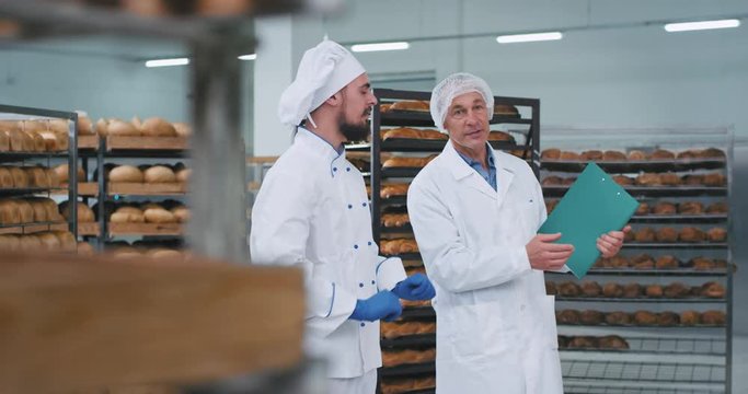 In a bakery factory young baker and old man engineer have a conversation engineer giving a instruction note for the baker background full shelves of baked fresh bread