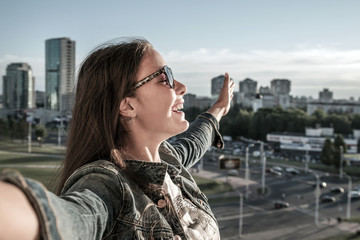 young brunette girl is standing on balcony with her hands spread wide, beautiful female happy teenager portrait in sunglasses and denim clothes, city background