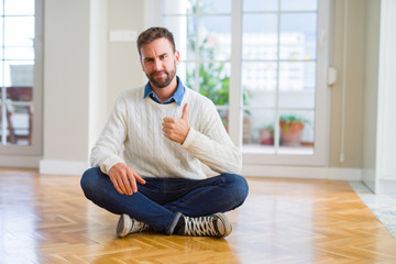 Handsome man wearing casual sweater sitting on the floor at home doing happy thumbs up gesture with hand. Approving expression looking at the camera with showing success.