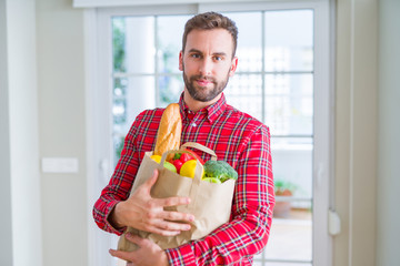 Handsome man holding groceries bag with a confident expression on smart face thinking serious