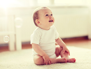 childhood, babyhood and people concept - happy little baby boy or girl sitting on floor with soap bubbles around at home