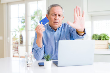 Handsome senior man shopping online using credit card and laptop at home with open hand doing stop sign with serious and confident expression, defense gesture