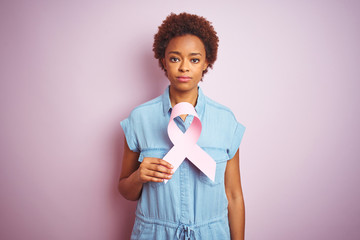 Young african american woman holding brest cancer ribbon over isolated pink background with a...