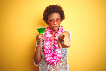Young african american woman with afro hair wearing flower hawaiian lei and drinking a cocktail pointing with finger to the camera and to you, hand sign, positive and confident gesture from the front