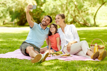 family, leisure and technology concept - happy mother, father and daughter having picnic and taking selfie by smartphone at summer park