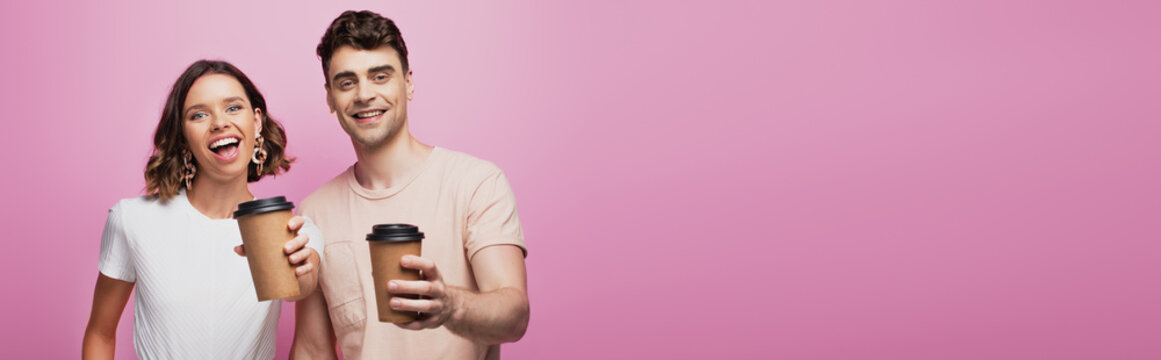 panoramic shot of happy man and woman hoding paper cups and smiling at camera on purple background