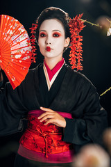 selective focus of beautiful geisha in black kimono with red flowers in hair holding hand fan and sakura branches isolated on black