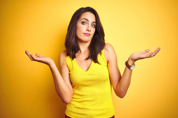 Young beautiful woman wearing t-shirt standing over yellow isolated background clueless and confused expression with arms and hands raised. Doubt concept.