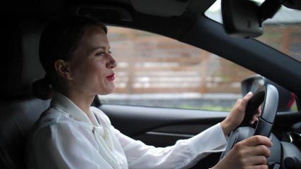 Close-up of smiling female chatting by phone in car using bluetooth hands free technology. Pretty woman in business clothes talking on in-car speakerphone while driving car through modern cottage town