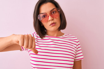 Young beautiful woman wearing fashion sunglasses with hearts over isolated pink background with angry face, negative sign showing dislike with thumbs down, rejection concept