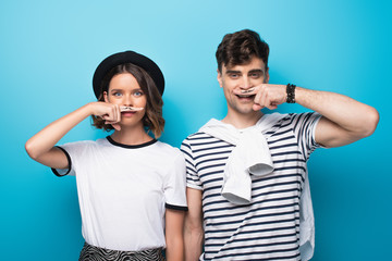 cheerful man and woman holding fingers with drawn mustache near faces on blue background