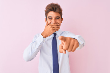 Young handsome businessman wearing shirt and tie standing over isolated pink background laughing at you, pointing finger to the camera with hand over mouth, shame expression
