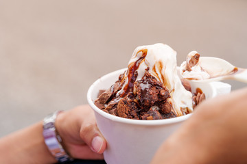 Outdoor street closeup view of hand hold a cup, plastic spoon, and eat melting frozen yogurt ice cream with chocolate topping in white paper cup.