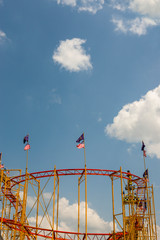 Achterbahn mit wehenden Fahne nach oben gegen den strahlenden und blauen Himmel mit Sonne und kleinen weißen Wolken fotografiert