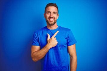 Young handsome man wearing casual t-shirt over blue isolated background cheerful with a smile of face pointing with hand and finger up to the side with happy and natural expression on face