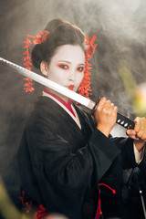 selective focus of geisha in black kimono holding katana in smoke and sakura branches