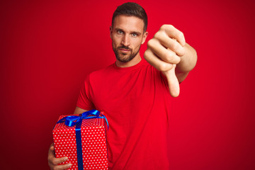 Young man holding birthday present over isolated red background with angry face, negative sign...