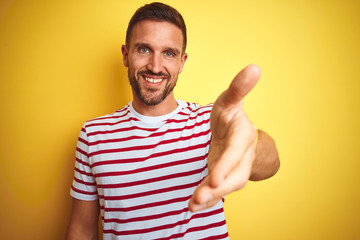 Young handsome man wearing casual red striped t-shirt over yellow isolated background smiling friendly offering handshake as greeting and welcoming. Successful business.